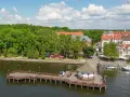 Malownicze wczasy na Mazurach: drewniany molo z altaną, jezioro, zielone drzewa i urokliwe miasteczko z czerwonymi dachami.