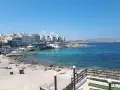 Słoneczny dzień na Bugibba beach. Ludzie odpoczywają na piasku i w turkusowej wodzie. W tle widać nadmorskie budynki.