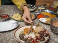 Ręka wybiera cynamon z talerza przypraw, przygotowując autentyczną kuchnię nepalską. Widać chili, mąkę, pastę imbirową i kolorowe przyprawy.