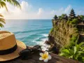 Słoneczny dzień na Bali. Słomkowy kapelusz, okulary i kwiat frangipani na drewnianym tarasie z widokiem na ocean i świątynię na klifie. Idealne miejsce na wakacje, po szczepieniach.