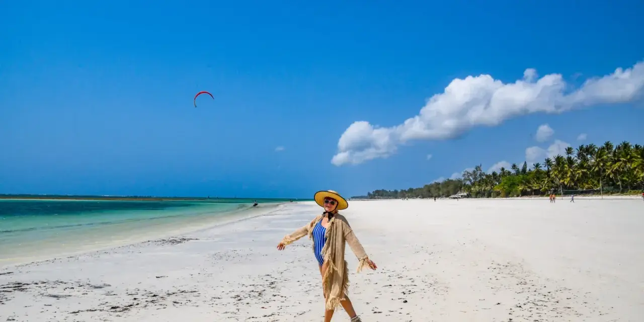 Kobieta w stroju kąpielowym i kapeluszu cieszy się słońcem na piaszczystej plaży Diani Beach. Na niebie czerwony latawiec do kitesurfingu.