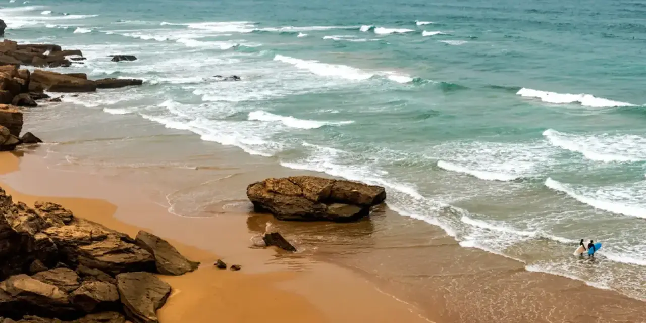 Maroko plaże: dwójka surferów wchodzi do wody z deskami, fale rozbijają się o skaliste wybrzeże.