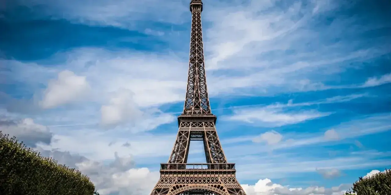 Majestatyczna wieża Eiffel w Paryżu, górująca nad zielonymi krzewami i błękitnym niebem z chmurami.