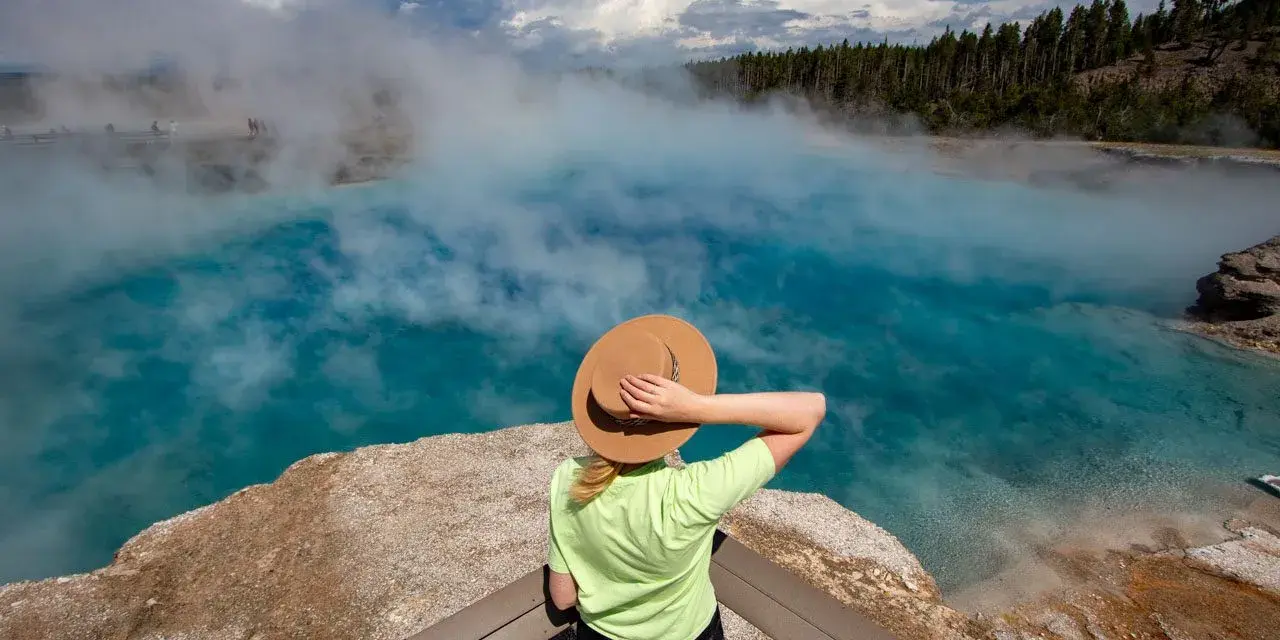 Kobieta w kapeluszu podziwia turkusowe gorące źródło w Yellowstone. Gdzie jest Yellowstone? Tu, wśród parujących wód i lasów.