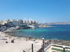 Słoneczny dzień na Bugibba beach. Ludzie odpoczywają na piasku i w turkusowej wodzie. W tle widać nadmorskie budynki.