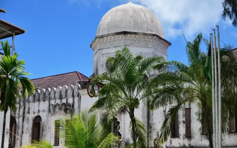 Biała, zabytkowa budowla z kopułą i palmami na tle błękitnego nieba. Klimatyczne zanzibar miasto.