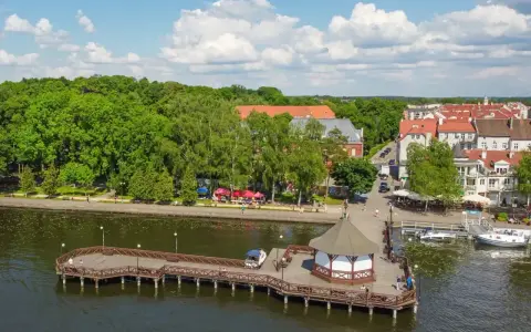 Malownicze wczasy na Mazurach: drewniany molo z altaną, jezioro, zielone drzewa i urokliwe miasteczko z czerwonymi dachami.