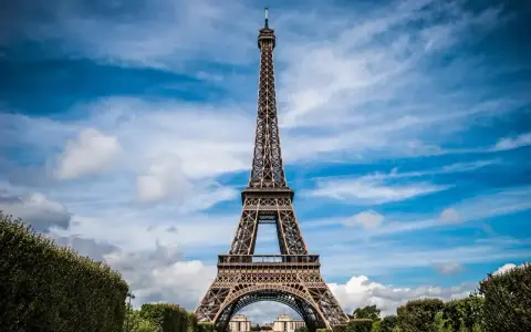Majestatyczna wieża Eiffel w Paryżu, górująca nad zielonymi krzewami i błękitnym niebem z chmurami.