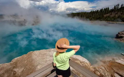 Kobieta w kapeluszu podziwia turkusowe gorące źródło w Yellowstone. Gdzie jest Yellowstone? Tu, wśród parujących wód i lasów.