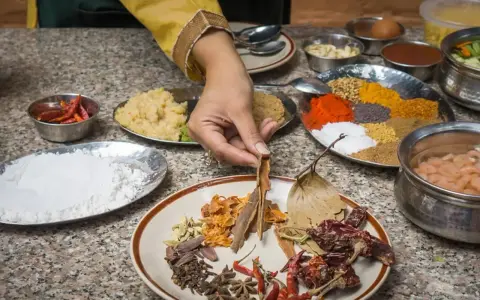 Ręka wybiera cynamon z talerza przypraw, przygotowując autentyczną kuchnię nepalską. Widać chili, mąkę, pastę imbirową i kolorowe przyprawy.
