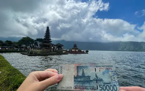 Ręka trzyma banknot 50 000 rupii na tle świątyni na wodzie. To symboliczne, że wiza turystyczna na Bali jest w zasięgu ręki!