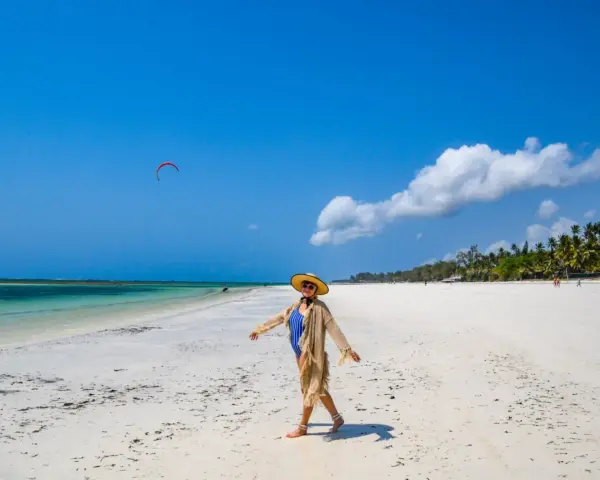 Kobieta w stroju kąpielowym i kapeluszu cieszy się słońcem na piaszczystej plaży Diani Beach. Na niebie czerwony latawiec do kitesurfingu.