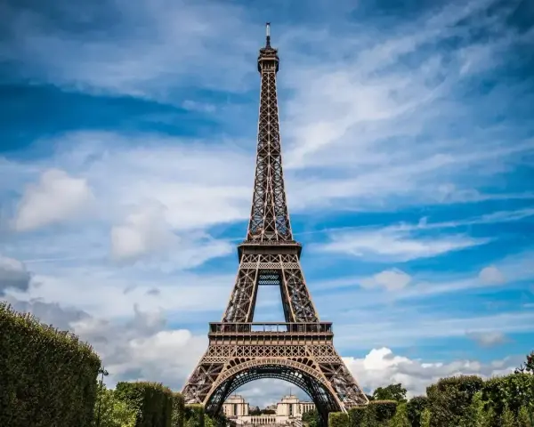 Majestatyczna wieża Eiffel w Paryżu, górująca nad zielonymi krzewami i błękitnym niebem z chmurami.