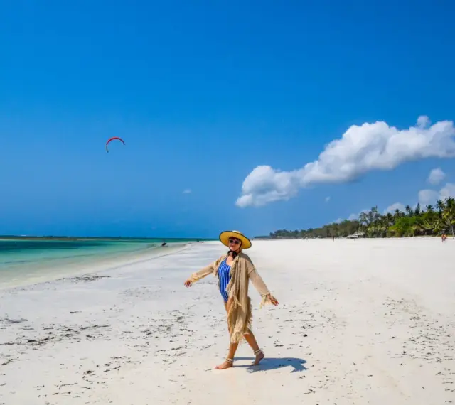 Kobieta w stroju kąpielowym i kapeluszu cieszy się słońcem na piaszczystej plaży Diani Beach. Na niebie czerwony latawiec do kitesurfingu.
