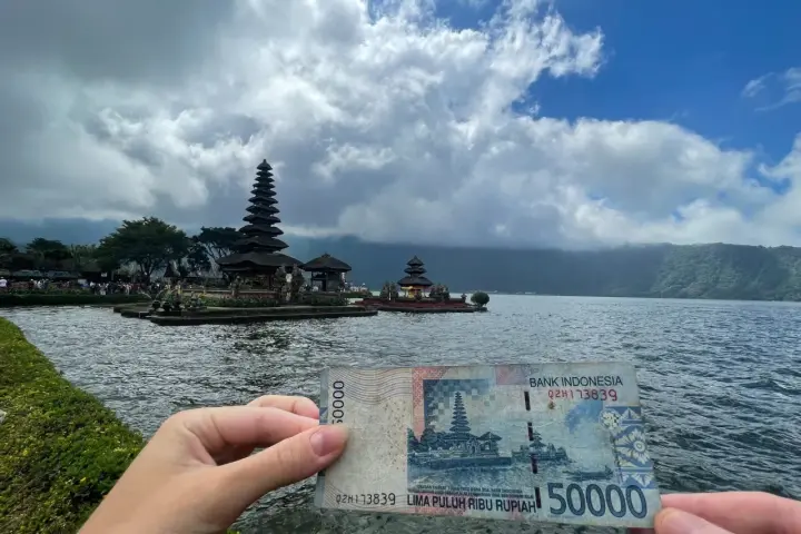 Ręka trzyma banknot 50 000 rupii na tle świątyni na wodzie. To symboliczne, że wiza turystyczna na Bali jest w zasięgu ręki!