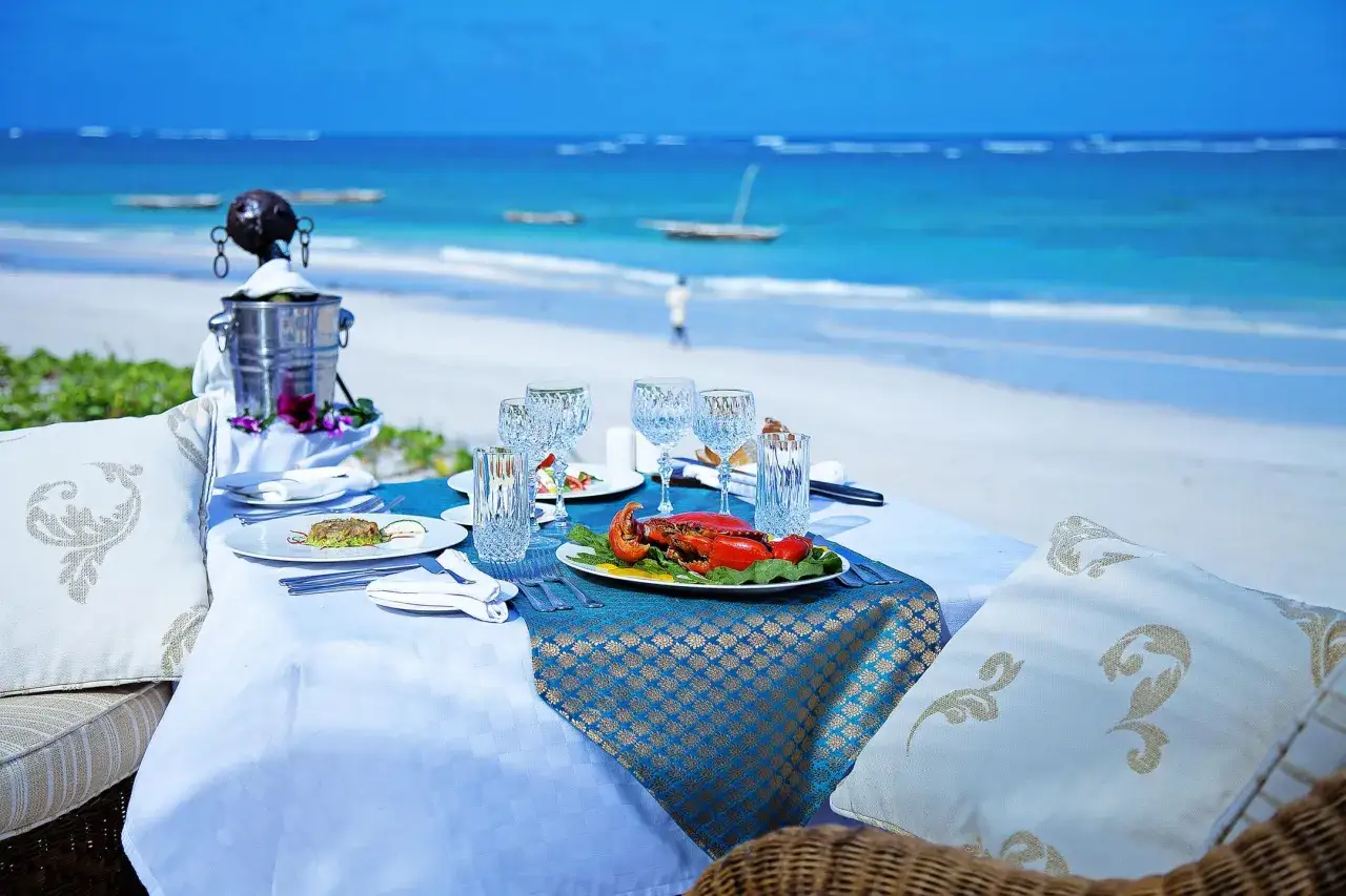 Romantyczna kolacja na plaży Diani: stół nakryty do posiłku z homarem, kieliszki, butelka szampana w wiaderku, w tle ocean i biały piasek.