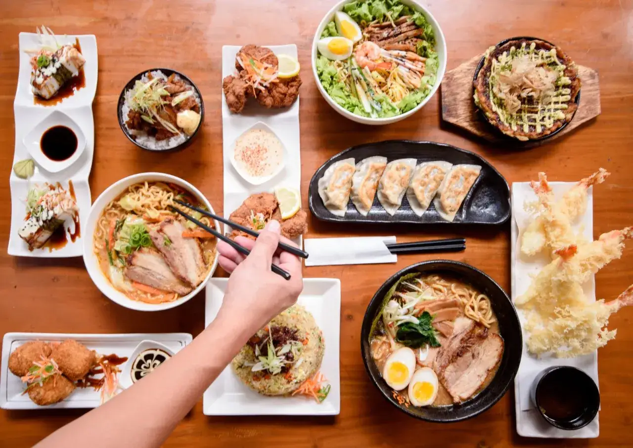 Obfity stół z japońskim jedzeniem: ramen, sushi, tempura, gyoza, kurczak karaage i sałatka. Ręka z pałeczkami sięga po miskę z ramenem.