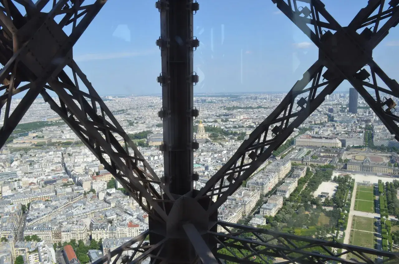 Widok na Paryż z wnętrza konstrukcji wieży eiffel. Widać dachy budynków i zieleń.