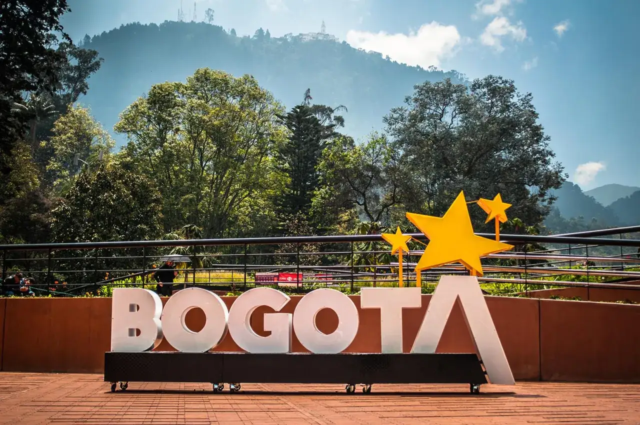 Napis BOGOTA z żółtymi gwiazdami, symbolizujący stolicę Kolumbii, na tle zielonych wzgórz i błękitnego nieba.