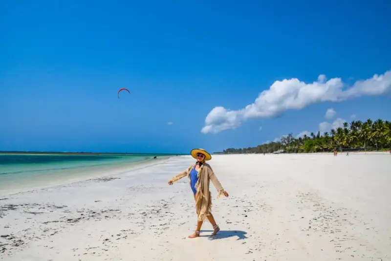 Kobieta w stroju kąpielowym i kapeluszu cieszy się słońcem na piaszczystej plaży Diani Beach. Na niebie czerwony latawiec do kitesurfingu.